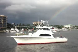 A white fishing boat with a red stripe floats on the water near a marina, with buildings and other boats in the background. A faint rainbow appears in the cloudy sky above.